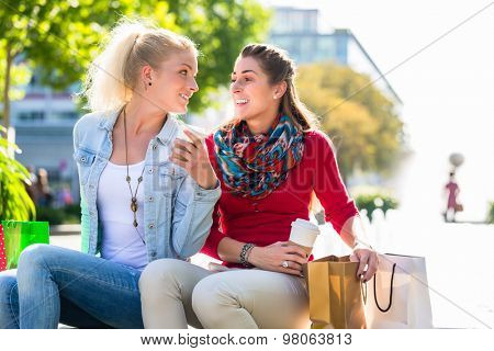 Two women friends sitting on fountain rim in city with shopping bags