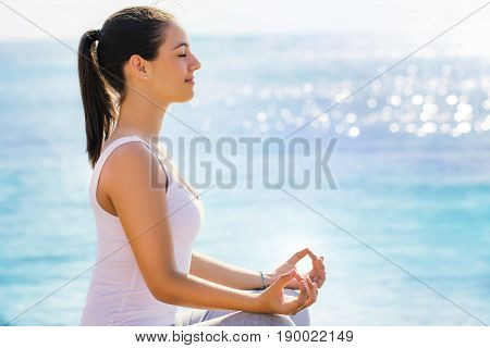 Close up side side view portrait of young woman in casual wear searching for spiritual enlightenment next to sea. Girl doing yoga exercise in early morning sun with bright reflection on water surface.