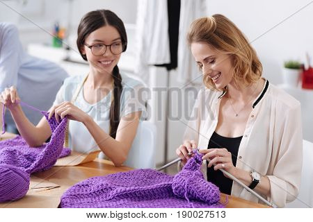 We love knitting. Two pleasant cheerful women sitting at the table and knitting scarves with purple threads and discussing something pleasant