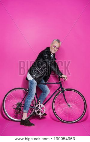 Happy Fashionable Senior Man Wearing Leather Jacket, Sunglesses And Riding Bicycle Isolated On Pink