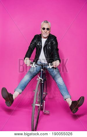 cheerful stylish senior man wearing leather jacket riding bicycle isolated on pink