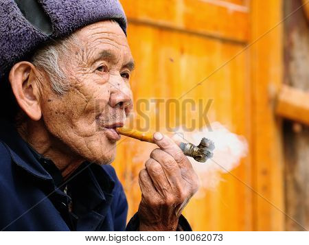 ZHAOXING CHINA - 11 NOVEMBER 2010: Old Chinese resting on the doorstep of his cottage smoking a pipe in the village Zhaoxing