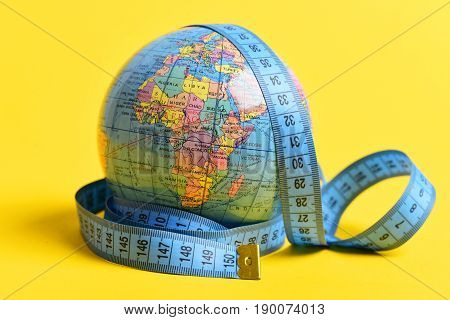 Tape for measuring wraps around geographical globe isolated on warm yellow background. Symbol of worldwide economic and social issues