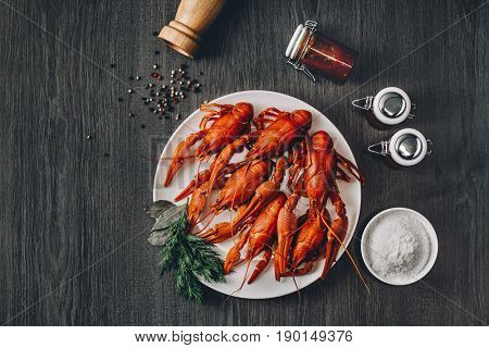 Boiled big red fresh crawfish in white plate with green herbs. Salt bowl, sauce jars and pepper grains around. Gray wooden background. Instagram vintage toning effect. Top view. Copy space.