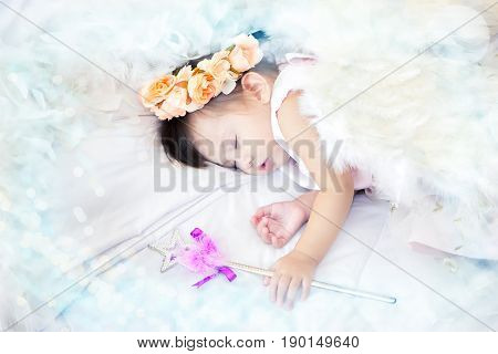A mestizo lovely girl dressed as a fairy has a crown pastel flowers on her head sleeping and magic scepter in her hand on the couch white and white feather place surrounded and copy space