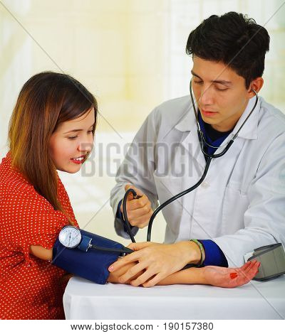 Close up of a handsome young doctor with stethoscope around his neck taking the pulse to a young woman with a tensiometer, in a doctor consulting room background.