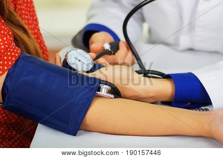 Close up of a young doctor using the stethoscope to hear the pulse while a woman is with a tensiometer in her arm, in a doctor consulting room background.
