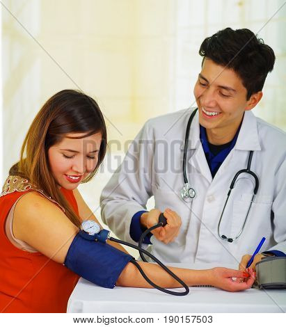 Close up of a handsome smiling young doctor with stethoscope around his neck taking the pulse to a young woman with a tensiometer, in a doctor consulting room background.