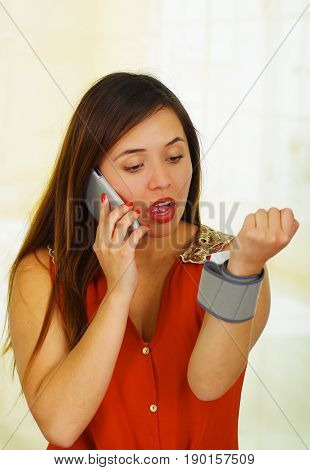 Beautiful young patient using an automatic tensiometer in her arm and calling in her cellphone, in a doctor consulting room background.