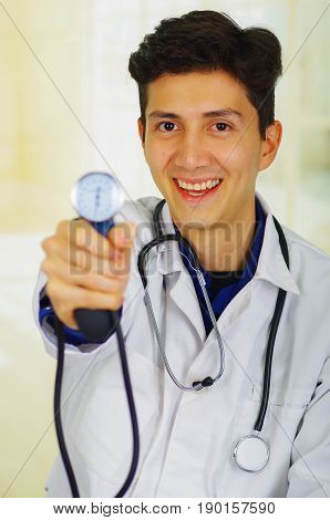 Handsome smiling young doctor with stethoscope around his neck, holdig a Tensiometer in his hand, in a doctor consulting room background.