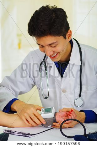 Handsome smiling young doctor with stethoscope around his neck taking the pulse to a young woman, in a doctor consulting room background.