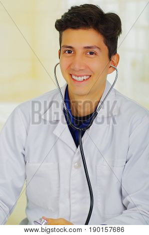 Handsome smiling young doctor with stethoscope around his neck, pposing for camera, in a doctor consulting room background.