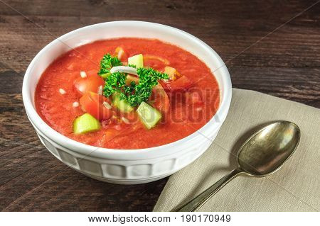 A bowl of gazpacho soup with slices of tomato, cucumber, and onion, with parsley leaves,  on a dark rustic texture with a place for text
