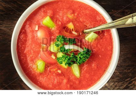 A closeup of gazpacho soup with slices of tomato, cucumber, and onion, with parsley leaves, shot from above on a dark rustic texture