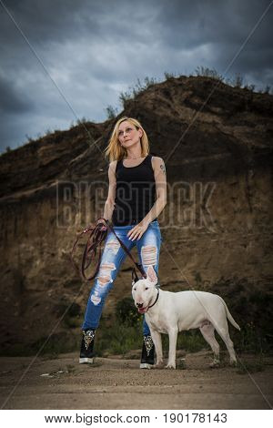 Brutal, bold girl with a fighting dog breed of bull terrier on a background of a rock in the evening