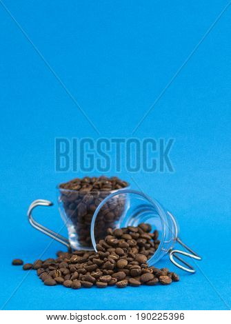 Two glass cups one standing full of coffee grains and the other leaning on the tanle spreading coffee grains. With a light blue background.
