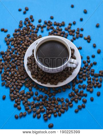 white cup full of coffee with coffee grains on the saucer and on a light background surrounding the cup we see coffee grains.