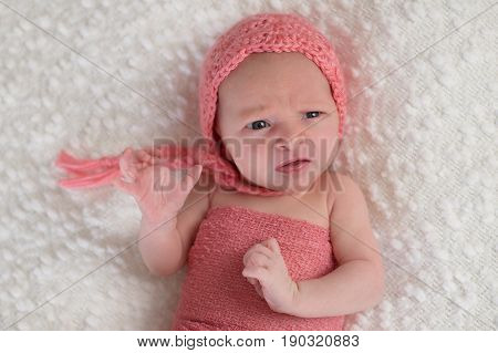 A three week old newborn baby girl wearing a peach colored bonnet. She is lying on a white bouncle blanket and has a funny expression on her face.