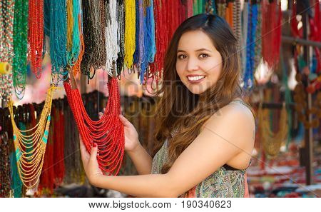 Beautiful caucasian woman holding a finished traditional necklace in her hands and smiling to the camera, handicrafts art, colorful necklace background.