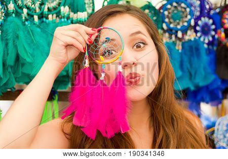 Close up of a beautiful smiling young woman pointing in from of her face a pink catchdreamers, in colorful market fabrics background.
