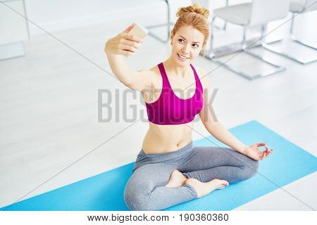 Portrait of fit red haired woman doing yoga exercises at home: taking selfie with smartphone sitting in lotus position