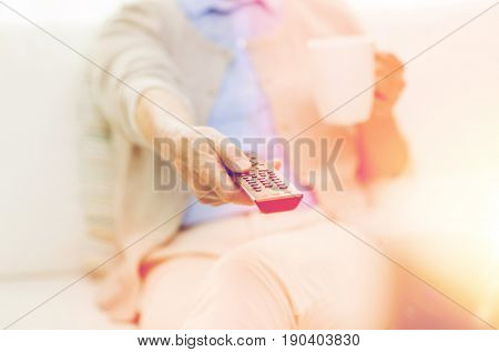 technology, television, age and people concept - close up of senior woman watching tv, drinking tea and changing channels by remote control at home