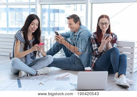 Modern igeneration. Delighted positive young people sitting together and holding their smartphones while using modern technology