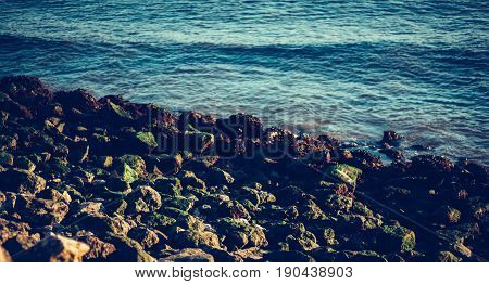 Close-up horizontal view of beautiful pebble coastline of beach with teal calm water stones with moss and dehydrated algae sunny summer day in Lisbon Portugal shallow depth of field