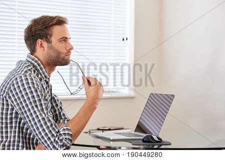 Handsome young man seated behind his laptop computer, looking into the distance in thought, concentrating on all the business decision she has to make in order to become a success.