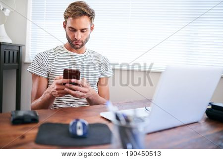 Good looking young caucasian guy busy using his phone with both hands while sitting at home behind his computer at his desk next to a nice large window.