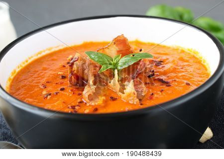 Thick tomato soup with basil and fried bacon in a black ceramic bowl on a grey abstract background. Healthy eating concept Fresh tomato soup with creme fraiche