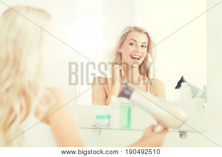 beauty, hairstyle, morning and people concept - smiling young woman with fan blow drying her hair looking to mirror at home bathroom