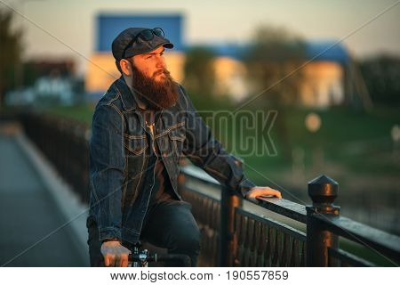Bearded guy and fix bike. Young man with gourgeous large beard in sunglasses and in the cap riding a vintage bicycle in the city park at sunset.
