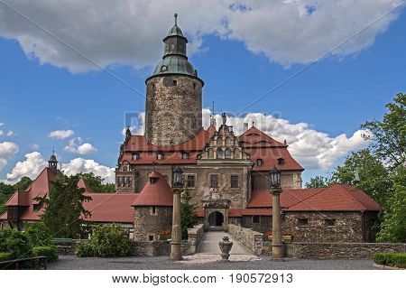 The photograph shows the historic Czocha castle located in Forest in south-west Poland. There is a sunny day, the sky is lightly clouded. Around the castle tall trees grow. He is leading a stone bridge to the castle.