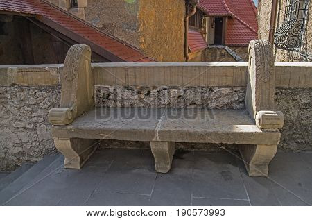 The photo shows an old stone bench. It stands beside a stone wall. At the back you will see the walls and roofs of the castle buildings. It is an architectural element of the castle of Czocha in the village of Lesna, in south-western Poland.