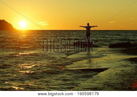 Silhouette of a man standing on a rock with raised perpendicular hands on a sunset background on the seashore.