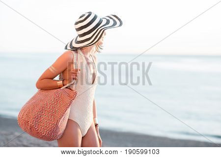 Young woman in hat and swimsuit walking with beach bag near the ocean during the summer vacation