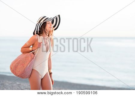 Young woman in hat and swimsuit walking with beach bag near the ocean during the summer vacation