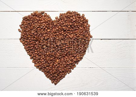 Pile of roasted brown coffee beans in form of heart shape over white wooden table, top view background
