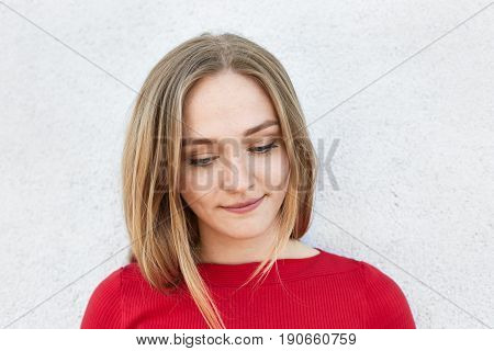 Sensual woman with fair hair and beautiful face looking down thoughtfully having some dreams in her mind while posing in white studio. Pensive woman with beautiful appealing appearance in red clothes
