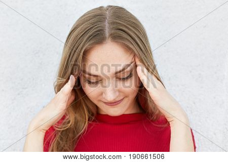 Indoor shot of tired fair-haired woman in red sweater holding her hands on temples having headache after hard work looking down. Woman wanting to remember some details having pensive look down