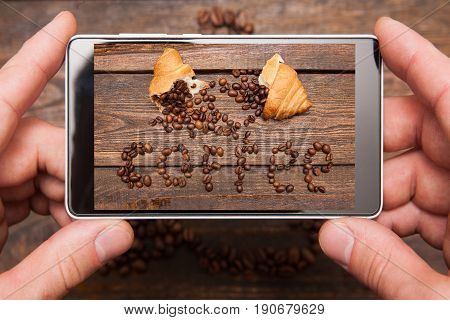 Mobile conceptual photography. Phone in hands taking photo of insctiption made by coffee beans and sweet dessert on wooden background for social network, top view