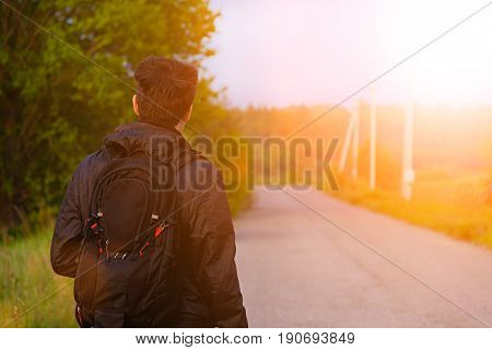 Young man with backpack walking on the road along forest at dawn away from the viewer