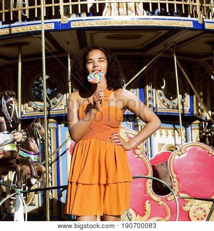 cool real modern teenage girl with candy near carousels at amusement park walking, having fun