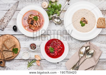 Set of three bowls with different soups: meat soup solyanka, Russian borsh and mushroom cream soup on white wooden background, top biew.