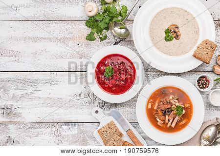 Set of three bowls with different soups: meat soup solyanka, Russian borsh and mushroom cream soup on white wooden background, top biew.