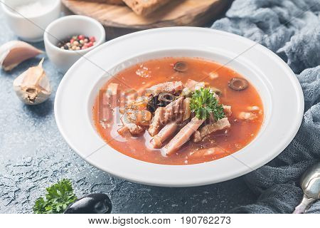Soup with meat, olives, herbs, lemon in white bowl, black bread and spices on gray background, homemade food. Traditional Russian soup - solyanka