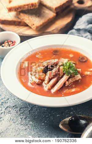 Soup with meat, olives, herbs, lemon in white bowl, black bread and spices on gray background, homemade food. Traditional Russian soup - solyanka