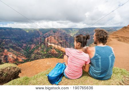 People hiking in Hawaii hikers pointing at Kauai. happy hiker couple healthy lifestyle outdoors looking at Waimea canyon view. Young couple resting sitting in nature in Kauai, Hawaii, USA.