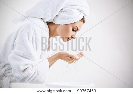 Young woman washing face with clean water in white bathrobe and towel. Morning hygiene concept.